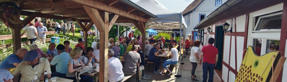 Bei strahlendem Sonnenschein sitzen die Besucher im großen Garten und Hof des „Selbstversorgerhauses Lützel“ beisammen und lauschen den Klängen von „BrützelBrass“. (Foto: DF)